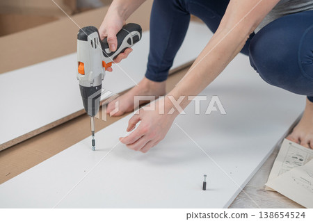 A Caucasian woman uses a screwdriver to tighten a screw while assembling a cabinet. A Caucasian woman uses a screwdriver to tighten a screw while assembling a cabinet. 138654524