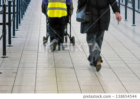 Passenger walking to airport departure gate, assistance staff helping with person on wheel chair, view from behind Passenger walking to airport departure gate, assistance staff helping with person on wheel chair, view from behind 138654544