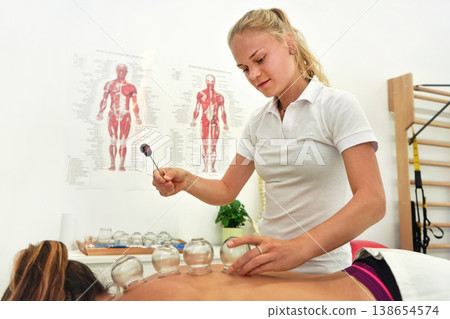 Young female physiotherapist applying glass suction banks on back of her patient, holding open flame above during cupping therapy, closeup detail Young female physiotherapist applying glass suction banks on back of her patient, holding open flame above during cupping therapy, closeup detail 138654574