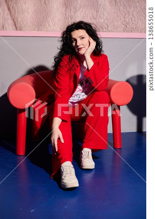 Cheerful woman dressed in red seated on pink sofa expressing amusement Cheerful woman dressed in red seated on pink sofa expressing amusement 138655018