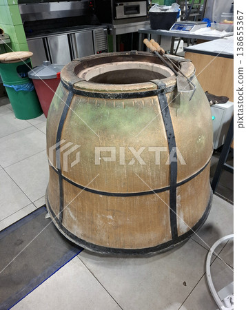 A large clay tandoor oven is placed in a professional kitchen, surrounded by cooking tools and equipment used for preparing various dishes. Smoke and heat are present in the air. A large clay tandoor oven is placed in a professional kitchen, surrounded by cooking tools and equipment used for preparing various dishes. Smoke and heat are present in the air. 138655367