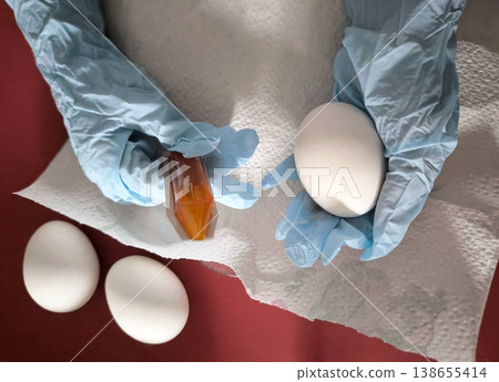 Child's hands coloring a white egg with food coloring while wearing blue protective gloves. Child's hands coloring a white egg with food coloring while wearing blue protective gloves. 138655414