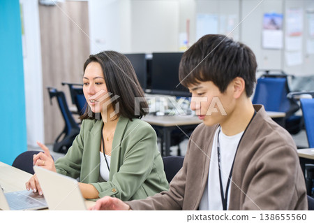A female senior colleague gives advice to a male employee who is working on his computer in the office. 138655560