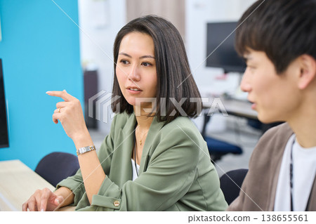 A female senior colleague gives advice to a male employee who is working on his computer in the office. A female senior colleague gives advice to a male employee who is working on his computer in the office. 138655561