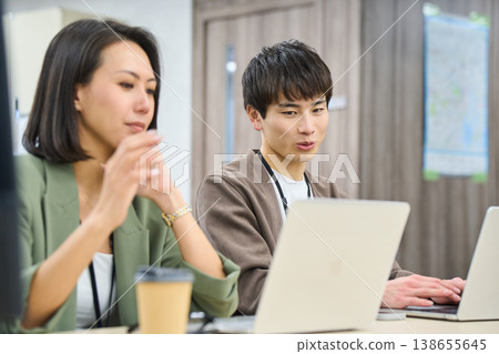 A young male employee works in a free-address office, consulting with a female senior colleague. 138655645