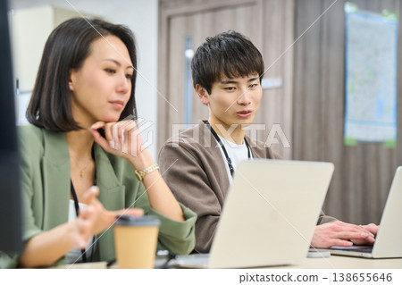 A young male employee works in a free-address office, consulting with a female senior colleague. A young male employee works in a free-address office, consulting with a female senior colleague. 138655646