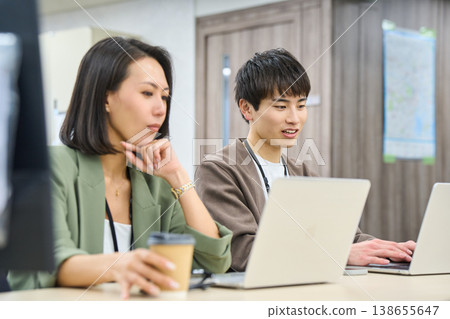 A young male employee works in a free-address office, consulting with a female senior colleague. A young male employee works in a free-address office, consulting with a female senior colleague. 138655647