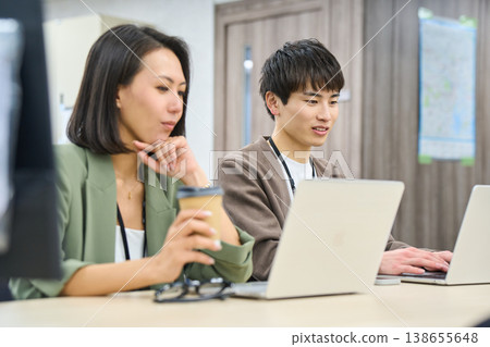 A young male employee works in a free-address office, consulting with a female senior colleague. A young male employee works in a free-address office, consulting with a female senior colleague. 138655648