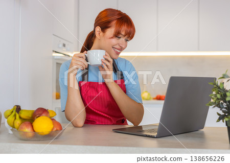 Young woman in kitchen with laptop answers emails writes replies and drinks coffee focused workflow 138656226