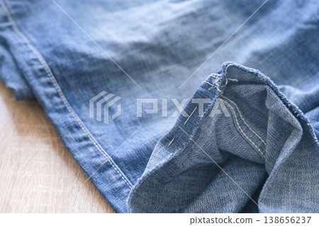 Blue denim fabric prepared for shortening or repair, with a single pin holding the folded hem in place. Denim pants hemmed for alterations with sewing pin Blue denim fabric prepared for shortening or repair, with a single pin holding the folded hem in place. Denim pants hemmed for alterations with sewing pin 138656237