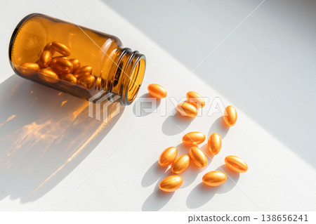 Amber bottle spilling golden softgel capsules on white background, representing medicine, vitamins, and skincare. Bottle spilling golden pills for health and beauty 138656241