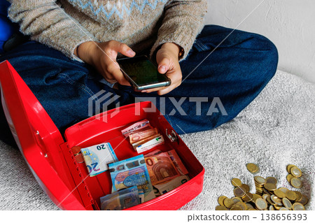 Young woman on bed using smartphone and counting euro cash from open cash box Young woman on bed using smartphone and counting euro cash from open cash box 138656393