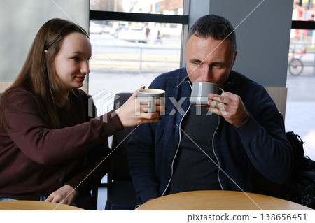 European father in blue sweater and 17-year-old daughter in brown sweater enjoy tea at a cafe. Cozy family moment in city. European father in blue sweater and 17-year-old daughter in brown sweater enjoy tea at a cafe. Cozy family moment in city. 138656451