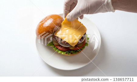 Hand wearing a white glove placing cheddar cheese on a hamburger with fast food hamburger preparation sandwich ingredient delicious appetizing restaurant barbecue homemade vegetables Hand wearing a white glove placing cheddar cheese on a hamburger with fast food hamburger preparation sandwich ingredient delicious appetizing restaurant barbecue homemade vegetables 138657711