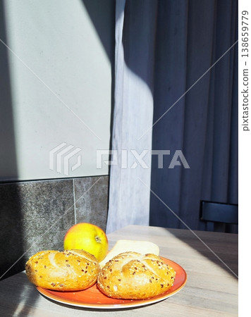 A sunlit wooden table holds a red plate with two round Kaiser bread rolls topped with seeds and an apple  138659779