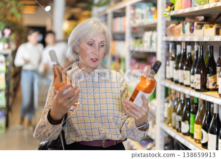 Senior female customer in supermarket examines bottles of wine, chooses alcoholic drink. Senior female customer in supermarket examines bottles of wine, chooses alcoholic drink. 138659913