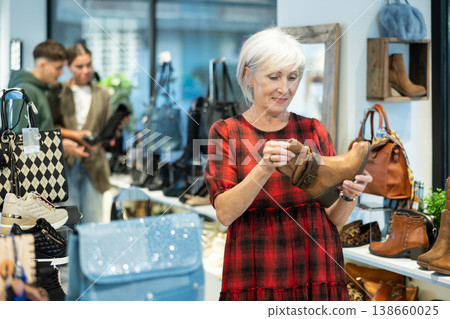 Elderly woman buyer looks at footwear from new collection and picks up new pair of boots for winter. 138660025
