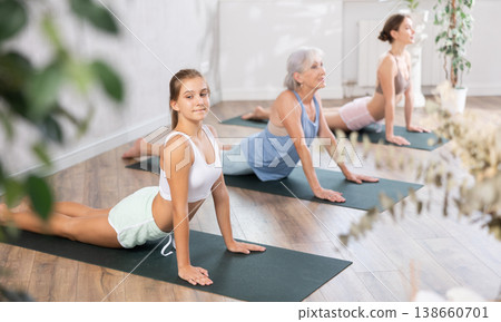 Women three generations practicing yoga positions during group training in fitness center, performing stretching asana Urdhva Mukha Shvanasana in studio Women three generations practicing yoga positions during group training in fitness center, performing stretching asana Urdhva Mukha Shvanasana in studio 138660701