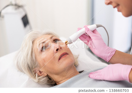 Young beautician doctor doing RF lifting procedure for face lifting of mature woman laying in beauty salon. Hardware cosmetology 138660703