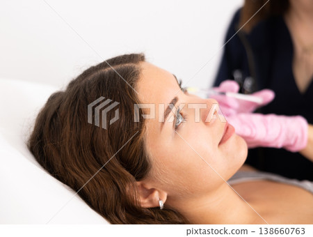 Hand of cosmetologist in gloves making rejuvenating injections procedure for tightening and smoothing wrinkles on face skin of young woman in beauty salon 138660763