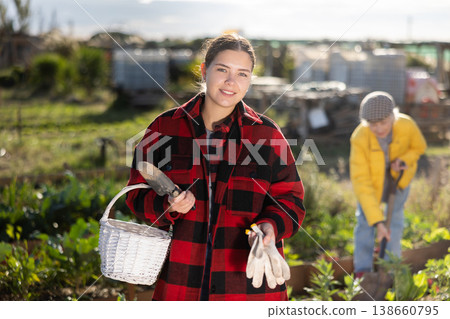 Smiling young female farmer posing with basket and garden instruments during season of sowing on sunny day of autumn Smiling young female farmer posing with basket and garden instruments during season of sowing on sunny day of autumn 138660795