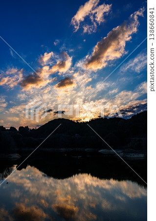 The setting sun paints the sunset sky in golden hues as it sinks behind the mountain range on the opposite shore of Lake Nunome, with golden clouds and reflections floating in the deep blue sky. The setting sun paints the sunset sky in golden hues as it sinks behind the mountain range on the opposite shore of Lake Nunome, with golden clouds and reflections floating in the deep blue sky. 138660841