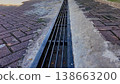 Metal storm drain grate on brick paved walkway. A long metal drainage grate runs through a brick-paved walkway and concrete gutter. The perspective shows parallel steel bars for water management in an 138663200