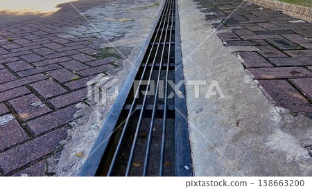 Metal storm drain grate on brick paved walkway. A long metal drainage grate runs through a brick-paved walkway and concrete gutter. The perspective shows parallel steel bars for water management in an Metal storm drain grate on brick paved walkway. A long metal drainage grate runs through a brick-paved walkway and concrete gutter. The perspective shows parallel steel bars for water management in an 138663200