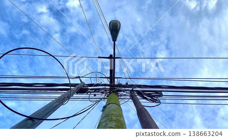 Tangled Utility Poles and Power Lines Against Blue Sky. A low-angle shot of weathered utility poles covered in moss with a complex network of electrical wires against a bright blue sky with light clou 138663204