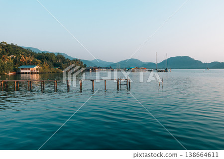 Long wooden pier stretching into calm sea towards a coastal fishing village. Long wooden pier stretching into calm sea towards a coastal fishing village. 138664611