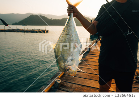 Fisherman holding a freshly caught fish on a wooden pier at sunset. 138664612