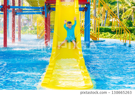 Caucasian cute happy kid boy rides water slide in a water park in summer vacation. Children's aqua park in pool at resort Caucasian cute happy kid boy rides water slide in a water park in summer vacation. Children's aqua park in pool at resort 138665263