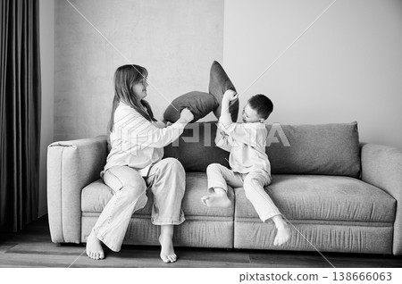Two young siblings started a pillow fight on couch. Little boy and her older sister fighting by pillows at home. Funny spending time with family. Black and white image. Two young siblings started a pillow fight on couch. Little boy and her older sister fighting by pillows at home. Funny spending time with family. Black and white image. 138666063