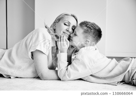 Little son showing love to his mom. Young mother and her kid lying on white carpet and spending time together at home. Lovely afternoon with family. Black and white image. Little son showing love to his mom. Young mother and her kid lying on white carpet and spending time together at home. Lovely afternoon with family. Black and white image. 138666064
