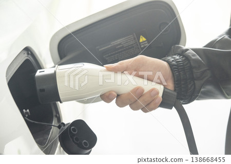 Hand inserting EV charging plug to electric vehicle with blurred background of outdoor natural greenery. Hand inserting EV charging plug to electric vehicle with blurred background of outdoor natural greenery. 138668455