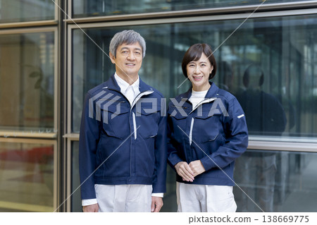 A man and woman in their 60s wearing work clothes smile and look directly at the camera. 138669775