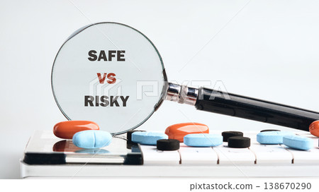 A magnifying glass reveals SAFE VS RISKY text prominently. Various orange, blue, and black pills lie scattered on a white calculating device A magnifying glass reveals SAFE VS RISKY text prominently. Various orange, blue, and black pills lie scattered on a white calculating device 138670290