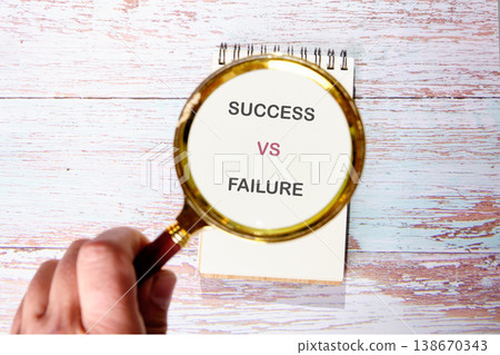 A hand holds a magnifying glass focusing on the text Success versus Failure written on a notebook resting on a light wooden surface 138670343