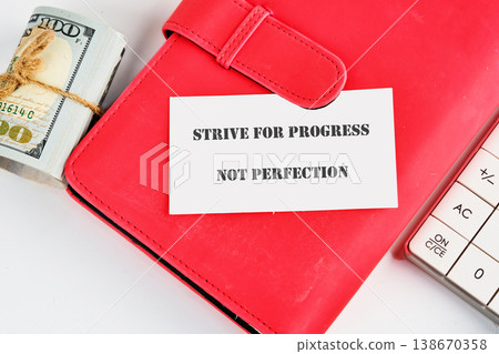 Red planner holds a card stating Strive for progress not perfection. Rolled US hundred dollar bills sit beside it, with a calculator on a white surface 138670358