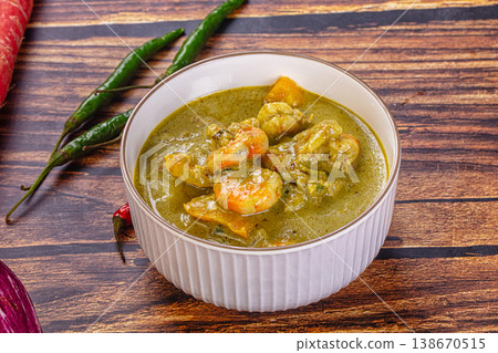 Thai green curry with shrimp and broccoli in a bowl, surrounded by fresh ingredients on a wooden table 138670515