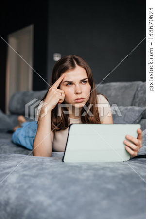 Young woman lies on the couch with a tablet in her hands and looks thoughtfully away. Young woman lies on the couch with a tablet in her hands and looks thoughtfully away. 138670623