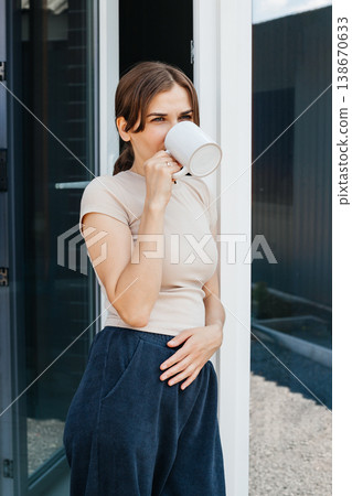 Young woman sips from a white mug, looking thoughtfully into the distance with a relaxed posture Young woman sips from a white mug, looking thoughtfully into the distance with a relaxed posture 138670633