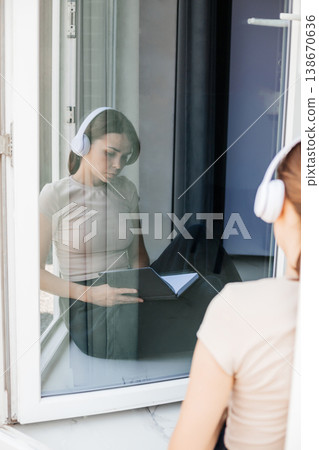 Young woman with headphones is sitting by the window and looking at her diary, you can see her reflection in the window. Young woman with headphones is sitting by the window and looking at her diary, you can see her reflection in the window. 138670636