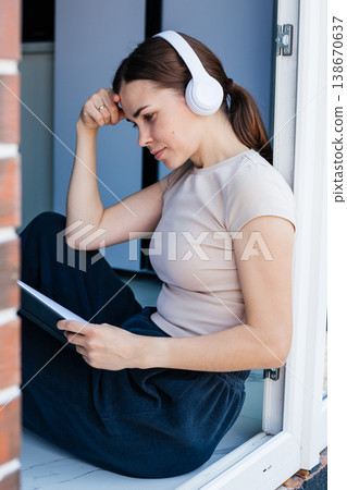 Young woman is sitting by the window wearing headphones and thoughtfully looking at her notebook. Student. Remote work. Young woman is sitting by the window wearing headphones and thoughtfully looking at her notebook. Student. Remote work. 138670637