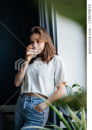 Young woman in casual clothes drinks water while standing by the window 138670654