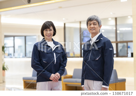 A man and woman in their 60s, wearing work clothes and smiling at the camera; they appear to be apartment building managers and facility staff. (Image) A man and woman in their 60s, wearing work clothes and smiling at the camera; they appear to be apartment building managers and facility staff. (Image) 138671017