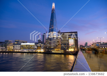 The Shard stands tall against the evening sky in London. People walk along the riverbank as lights from buildings reflect on the water. Construction cranes can be seen nearby. 138673682