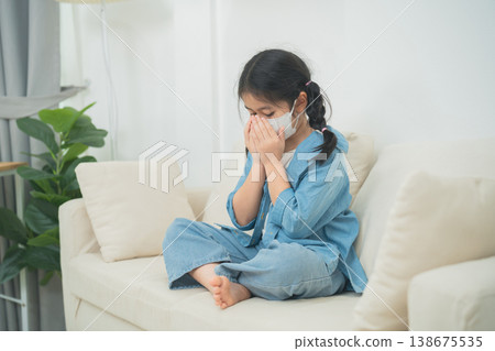 Young girl sitting on couch wearing mask, looking worried and frustrated, indoor setting with soft natural light and simple decor, family life and health concept 138675535