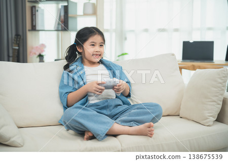 Happy young girl playing video game on couch in cozy living room, enjoying leisure time, lively expression reflecting excitement and fun Happy young girl playing video game on couch in cozy living room, enjoying leisure time, lively expression reflecting excitement and fun 138675539