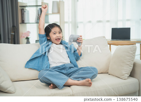 Joyful Girl Celebrating Victory on Couch While Playing Video Game, Wearing Casual Blue Outfit with Excitement and Happiness in Modern Living Room Setting 138675540